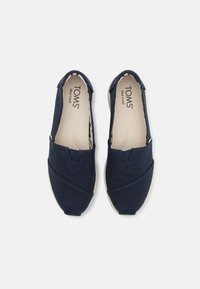 Marineblå kanvas slip-on sko med teksturert overflate, med avrundet tå, krysstikksdesign og lett gummisåle. TOMS-merkeinnvendig.