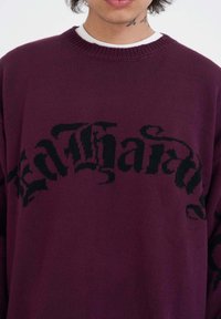 Burgundská pletená svetr s výrazným černým nápisem přes hruď. Kulatý výstřih se strukturovaným povrchem a dlouhými rukávy.