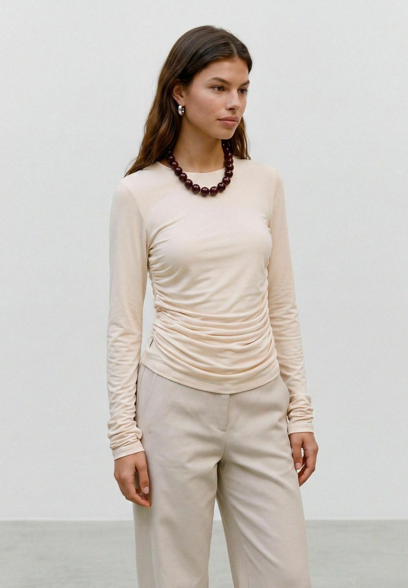 Femme aux longs cheveux bruns porte un haut beige à manches longues froncées, un pantalon assorti, un collier de perles foncé et des boucles d'oreilles argentées en forme d’anneau, debout devant un mur uni.