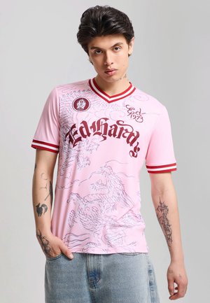 Giovane uomo con capelli corti e scuri che indossa una t-shirt rosa con grafica, bordi rossi e jeans blu, in piedi davanti a uno sfondo semplice.