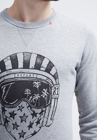 Sudadera gris con un gran gráfico negro de un casco con estrellas, gafas de sol que reflejan palmeras y el texto "REPLAY". Acento de costura roja.