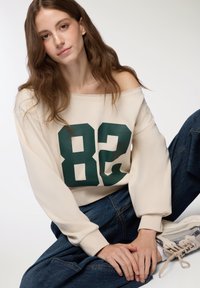 Felpa beige senza spalle con grande stampa verde '82', abbinata a jeans larghi blu scuro e sneakers chiare.