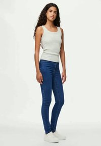 Ανοιχτό γκρι αμάνικο μπλουζάκι, μπλε στενά τζιν, λευκά αθλητικά παπούτσια. Το αμάνικο μπλουζάκι είναι εφαρμοστό με στρογγυλή λαιμόκοψη, ενώ τα τζιν είναι λεία με ελαφριά ελαστικότητα.