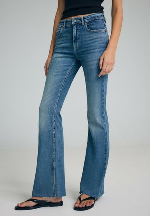 Kobieta w niebieskich dżinsach bootcut z surowymi szwami i czarnych klapkach stojąca na jednolitym tle.