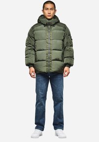 Olivgrüner Pufferjacke mit Kapuze, ausgestattet mit einem Reißverschluss auf der Vorderseite, vertikalen Nähten und elastischen Bündchen, getragen mit blauen Jeans und weißen Sneakers.