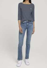 Top a righe blu e bianche con maniche a tre quarti indossato con jeans azzurri. Modella in piedi con sneakers bianche, mani ai lati, sfondo neutro.