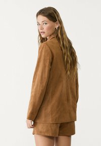 Junge Frau mit langen, welligen Haaren, die ein braunes Wildleder-Blazer und passende Shorts trägt und über ihre Schulter gegen einen schlichten Hintergrund schaut.