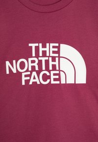 Bordové bavlněné tričko s velkým bílým grafickým prvkem a textem "THE NORTH FACE" a zakřiveným designem loga. Měkká textura.