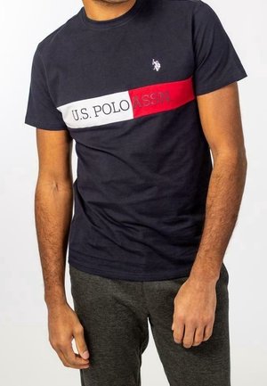 Mężczyzna ubrany w granatowy T-shirt U.S. Polo Assn. z czerwono-białymi poziomymi paskami oraz ciemnoszare spodnie na jednolitym tle.