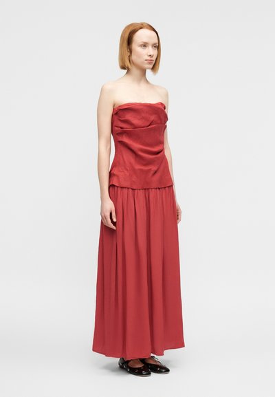 Φόρεμα χωρίς τιράντες, μακρύ έως το πάτωμα σε βαθύ κόκκινο χρώμα, με ρυτιδωτή υφή στο μπούστο και φαρδύ, ρέον μήκος. Συνδυάζεται με μαύρα παπούτσια φλατ.