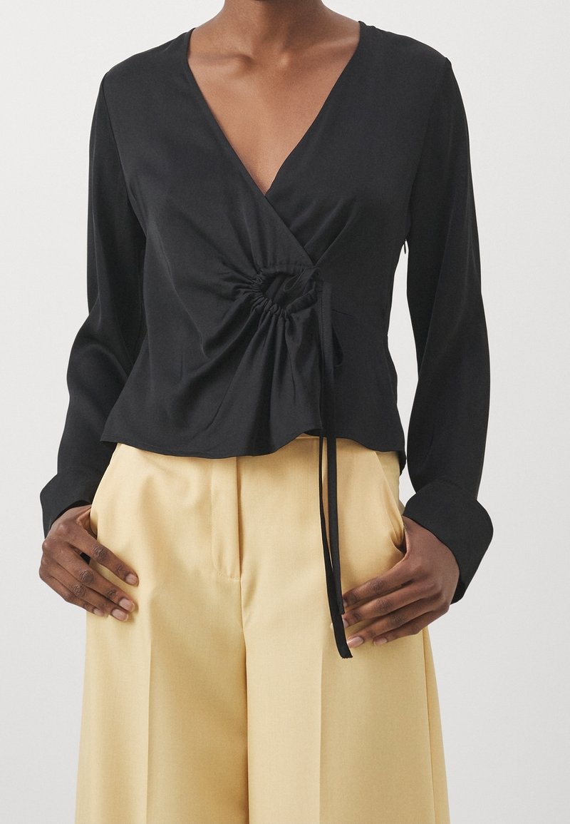 Femme portant un top cache-cœur à manches longues noir avec un détail froncé sur le côté et un cordon, associé à un pantalon large beige taille haute.