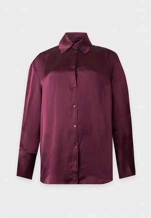 Donna in piedi con una camicia larga e lucida color marsala con bottoni, jeans blu e scarpe nere con punta appuntita su uno sfondo bianco semplice.