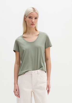 Blondynka w jasnozielonym krótkim rękawie oraz kremowych, wysokich spodniach, stojąca na tle jednolitego białego tła.