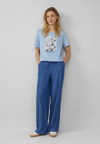 T-shirt bleu clair avec une impression graphique d'un paysage grec et le texte "Grèce", associé à un pantalon bleu ample et texturé et des chaussures décontractées.