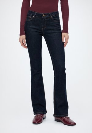 Femme portant un jean bootcut bleu foncé, un top rouge à manches longues et des chaussures en cuir rouges avec un détail chaîne argentée, debout sur fond blanc.