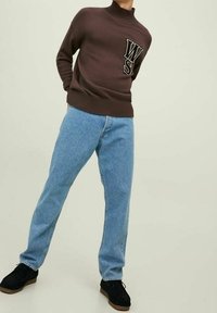 Bruine gebreide trui met hoge kraag en "WS" logo, gecombineerd met lichtblauwe denim jeans en zwarte sneakers met een tan zool.