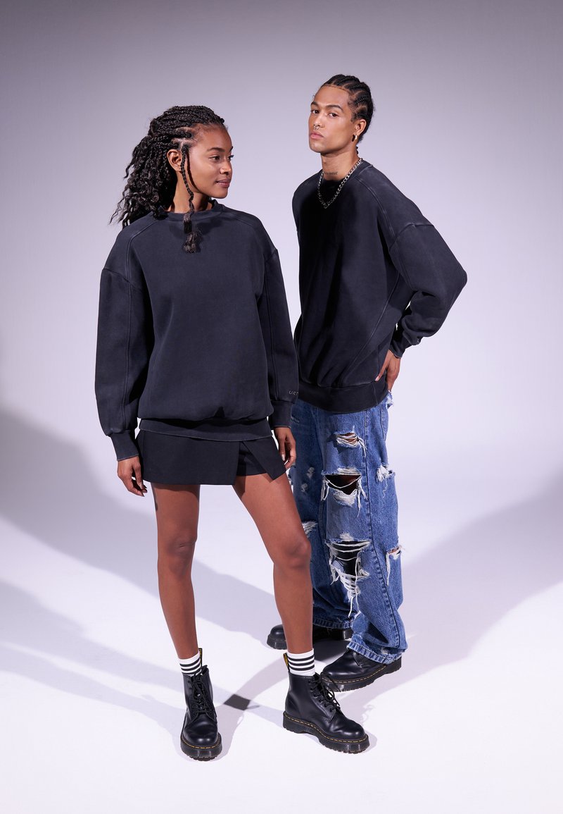 Dos modelos visten sudaderas negras de gran tamaño, una con una falda corta y botas negras, y la otra en jeans azules desgastados y botas negras.