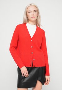 Jovem mulher a usar um cardigan vermelho brilhante com botões sobre uma blusa branca e uma saia de pele preta com uma bainha assimétrica.