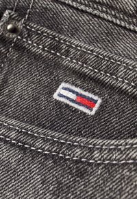 Mörkgrå denimtyg med en liten broderad logotyp i blått, vitt och rött. Sömnadsdetaljer synliga längs kanterna.