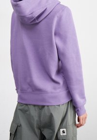 Lila huvtröja med ribbat nederkant och ärmar, tillverkad av ett mjukt tyg, bärs över gröna cargobyxor med en logotypapplikation.