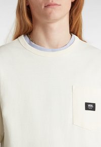 Camiseta de algodón color crema con un bolsillo en el pecho que lleva una etiqueta negra del logo de Vans, superpuesta sobre un cuello azul claro. Textura suave.