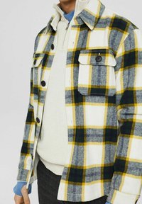 Flanellhemd in Weiß mit grün, gelb und grau kariertem Muster. Verfügt über schwarze Knöpfe, zwei Brusttaschen und einen umgeschlagenen Kragen.