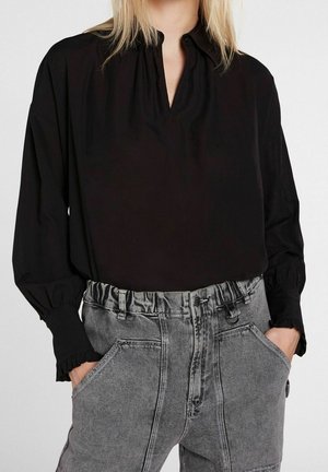 Blusa nera a maniche lunghe con scollo a goccia e polsini arricciati, abbinata a pantaloni di denim grigi a vita alta con grandi tasche.