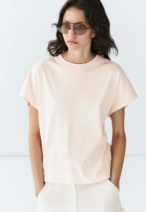 Femme aux cheveux foncés et ondulés portant un haut rose clair à manches courtes, un pantalon écru et des lunettes de soleil teintées marron, debout les mains dans les poches.
