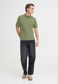 Polo vert olive à manches courtes avec col, associé à un pantalon noir ample et des baskets grises à lacets blancs.