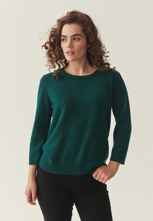 Woman with curly hair wears a dark green long-sleeve sweater and black pants, looking slightly to her left against a plain background.