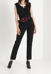 Tuta nera con scollatura profonda, caratterizzata da pieghe, abbinata a una cintura con borsa a tracolla in stampa pitone rossa e nera e stivaletti neri a punta.