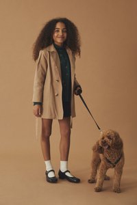 Beige dobbeltradet trenchcoat, grønn blomstrete skjorte, svart skjørt, hvite sokker, svarte Mary Jane-sko. Brun krøllete hund i bånd.