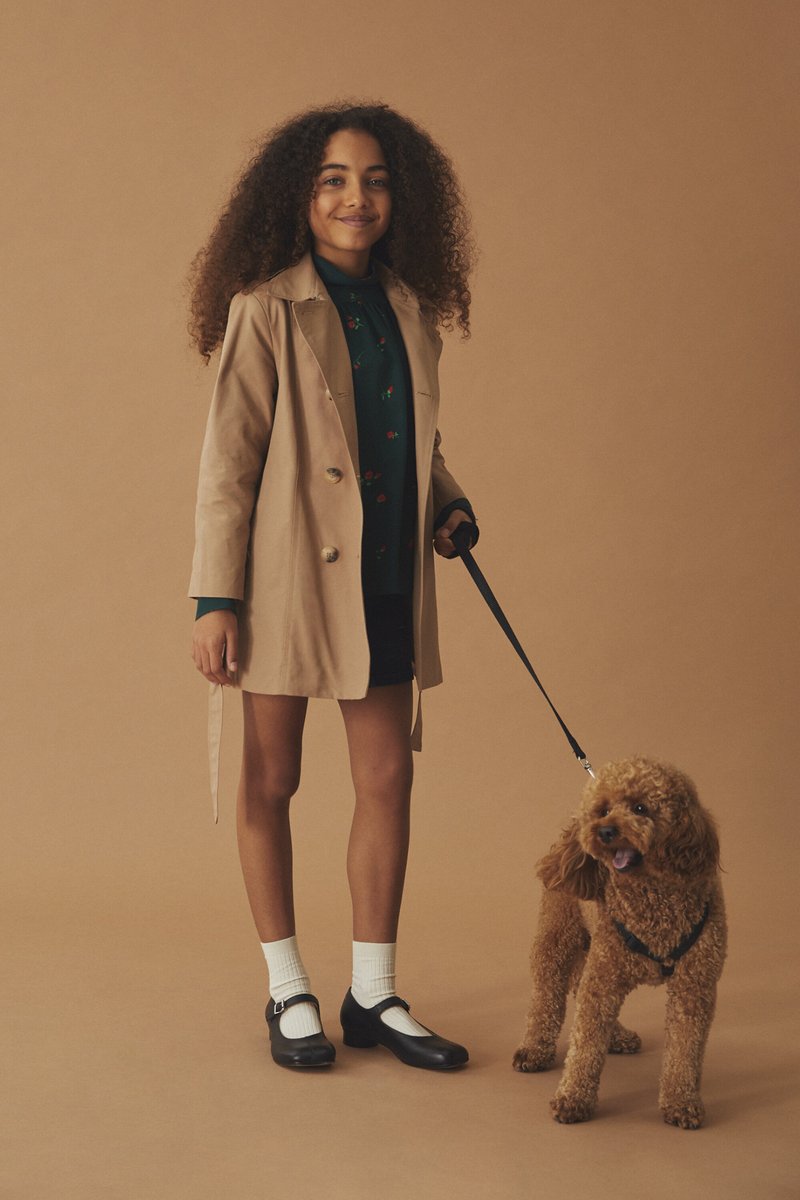 Beige dobbeltradet trenchcoat, grønn blomstrete skjorte, svart skjørt, hvite sokker, svarte Mary Jane-sko. Brun krøllete hund i bånd.