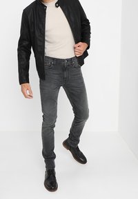 Černá kožená bunda přes béžovou košili, doplněná tmavě šedými slim-fit džíny a černými šněrovacími botami. Hladké textury a přiléhavý design.