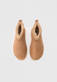 Ein Paar beige Wildleder-Ankleboots mit plüschigem Fellfutter und dekorativer Stickerei, von oben auf einem hellen Hintergrund betrachtet.