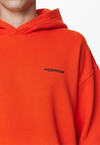 Hanorac portocaliu din material texturat, având pe față un logo negru "PEGADOR". Glugă cu sireturi, croi standard.