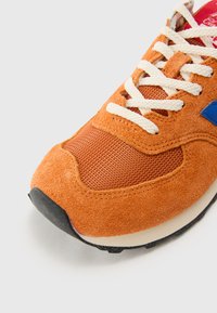Orange suede and mesh sneaker featuring white laces, blue accents, and black rubber sole. Textured surface with visible stitching details.