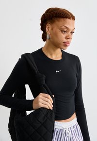 Svart langermet skjorte med rund hals og hvit Nike-logo, kombinert med en quiltet svart veske og bukser med hvite striper.