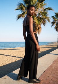 Woman in black crop top and wide pants standing on beach walkway, palm trees and ocean visible under clear blue sky.