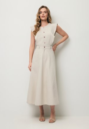 Vrouw met golvend haar, gekleed in een mouwloze beige midi-jurk met knopen en beige open-teen hakken, staand tegen een effen lichte achtergrond.