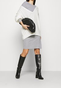 Pull blanc surdimensionné, robe grise à côtes, pochette noire texturée et bottes en cuir noir hautes, sur un fond uni.