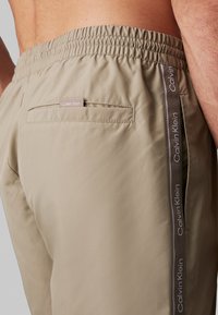 Boxer da bagno beige con cintura elastica, tasche laterali e etichetta di marca. Presenta una striscia logo a contrasto lungo i lati. Texture liscia.
