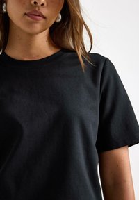 Czarna krótka koszulka bawełniana z okrągłym dekoltem; gładka tekstura i minimalistyczny design. Nie posiada widocznych logo ani wzorów.
