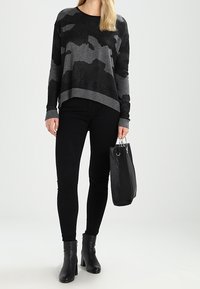 Černý a šedý maskáčový svetr s boxy střihem, žebrovaným lemem a manžetami, doplněný o černé úzké džíny a kotníkové boty; drží černou kabelku.