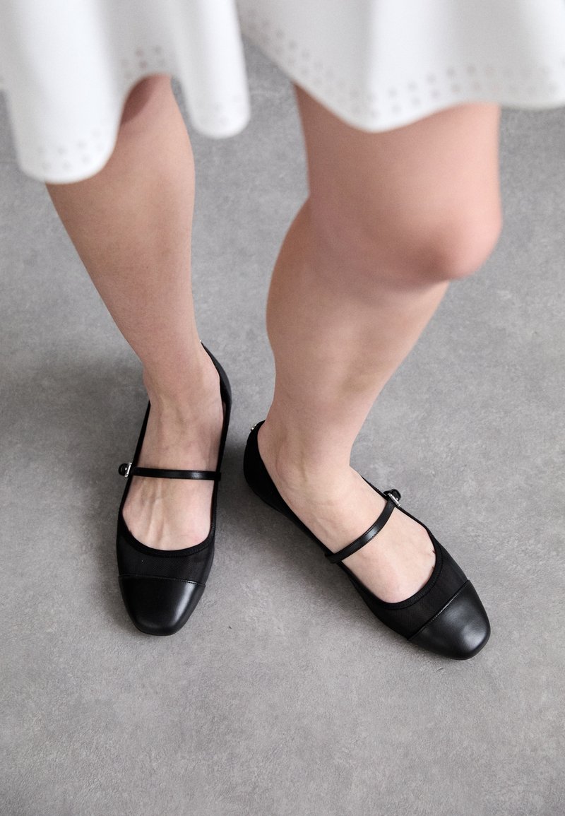 Schwarze flache Schuhe mit glänzendem Kappenbereich und Mesh-Obermaterial, versehen mit einem dekorativen Riemen über dem Spann und einer kleinen silbernen Schnalle.