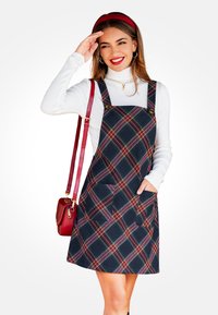 Vestido de cuadros oscuros con acentos en rojo y verde, con tirantes ajustables, dos bolsillos frontales y un diseño recto, combinado con un cuello de tortuga blanco.