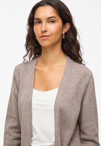 Ljusbrun cardigan i mjukt material med öppen framsida. Modellen bär en vit topp under.