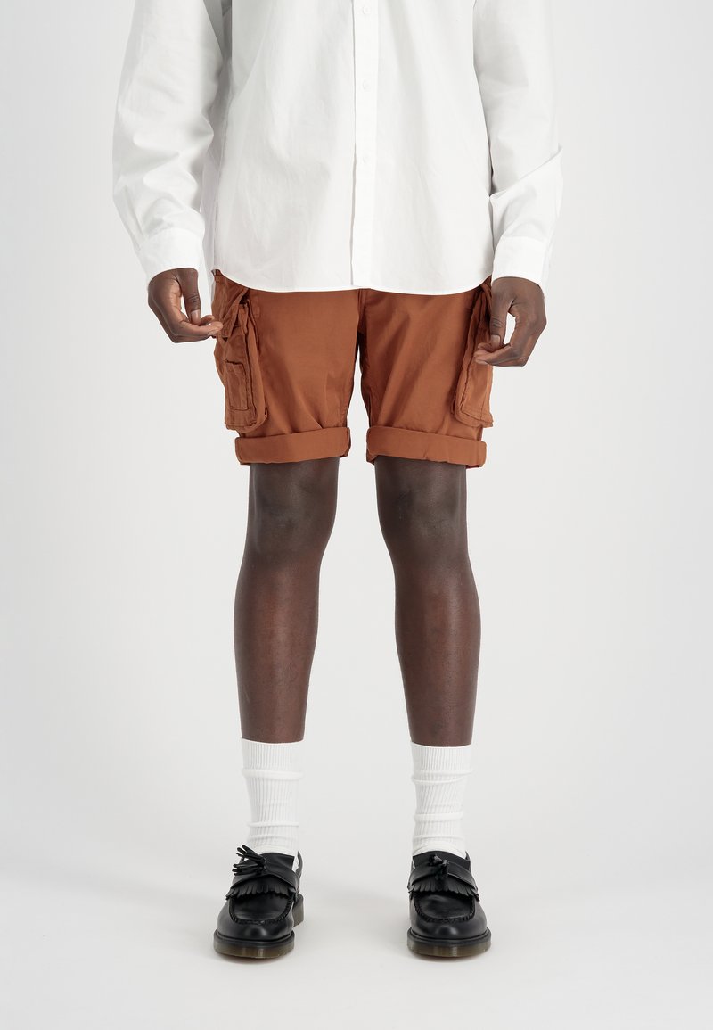 Person wearing rust-colored rolled-up cargo shorts, white long-sleeve shirt, white socks, and black tassel loafers standing on white background.