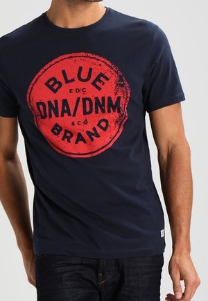 Mężczyzna w granatowej koszulce z dużym czerwonym okrągłym logo z napisem "BLUE BRAND DNA/DNM" w postarzałym stylu, do tego ciemne dżinsy.