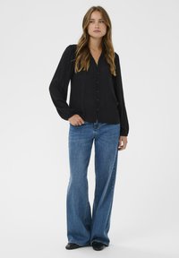 Sort langærmet bluse med V-hals, med knapper ned langs forsiden, parret med lyseblå flagrende jeans og sorte sko.